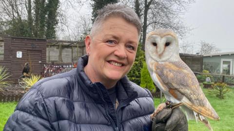 Sharon Cox, who has short white hair and is wearing a black puffer jacket, smiles at the camera. A small owl is perched on her gloved left hand and also looking towards camera. Over Sharon's shoulder a large black bird of prey is stood on a metal fixture in front of an aviary. 