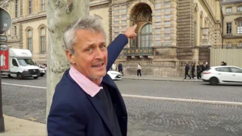 A man in a navy suit, looking to camera, pointing at a window in an old building behind him.