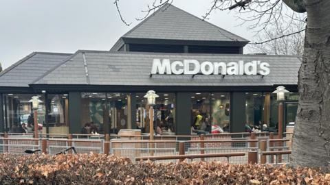 A one-storey building is in the middle of the image. There is a a hedge in front of the building and a tree on the right. A sign above the large windows says "McDonald's".