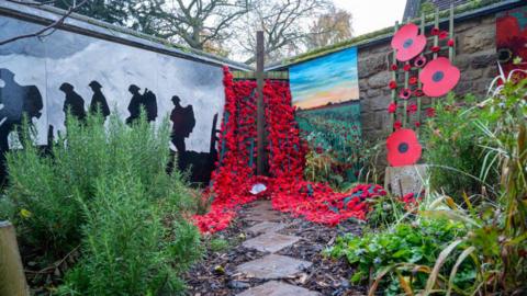 A garden with a stone path through the middle and a poppy display at the end mark with a large wooden cross. Another poppy display is to the right, while soldiers in silhouette are to the left.