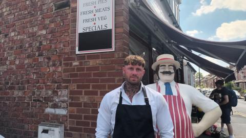 A younger man with tattoo on this neck, wearing a white shirt and a black apron, stands outside a red-brick butchers shop. A model butcher, with a hat, striped red apron and extravagant moustache, stands behind him.