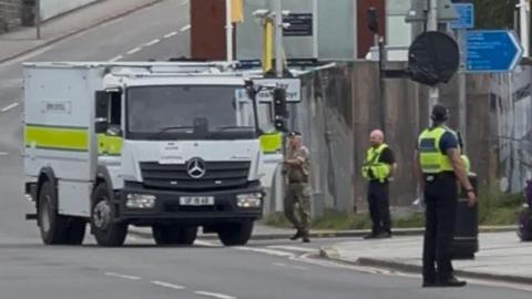 A large white truck on a street in Bridgend. It has a yellow stripe and man in military fatigues standing at the door. Two police officers are also on the street