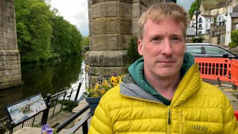 A man wearing a yellow coat stands in front of a collapsed walkway by the river.