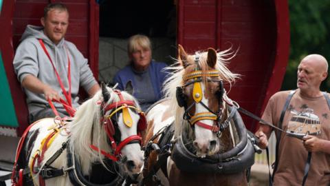 Fair-goers arrive in Cumbria, ahead of the Appleby Horse Fair, the annual gathering of gypsies and travellers. There are three people and two horses in bright yellow and red reins. The picture is from the Fair in 2023.