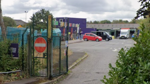 An entrance to a recycling centre with a metal gate, surrounded by hedges and a red car and two vans parked in front of large purple blocks behind that