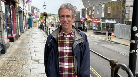 A man with grey hair wearing a black, white, and yellow plaid shirt and dark blue rain jacket with Market Jew Street in the background. 