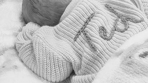 A close-up black-and-white image shows a small infant wrapped in a soft, knitted blanket. The blanket features thick ribbed textures and decorative stitched lettering across the fabric. Only part of the child's head is visible.
