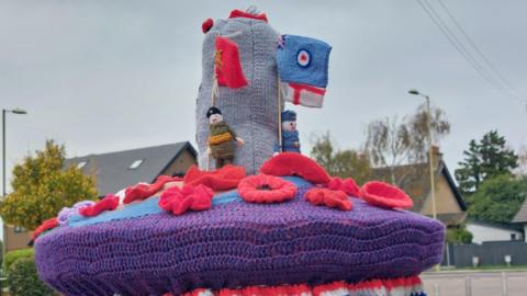 A postbox topper knitted with poppies, a solider and a sailor and the Royal Navy flag. They are standing next to a knitted cenotaph and behind there is a row of houses. The scene is on a residential street with houses behind.