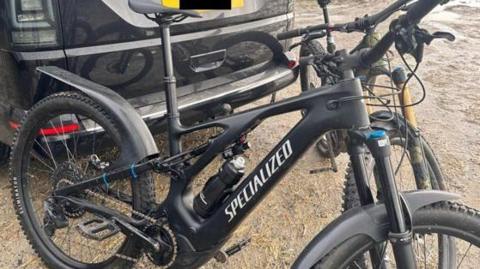 A black and silver electric mountain bike next to a green and black electric mountain bike, both made by Specialized, leaning next to a van