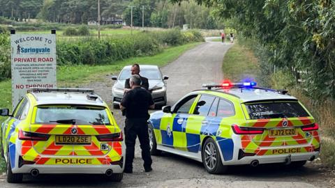 Two marked police cars close a private road near the event. Two officers can be seen placing cones in the road