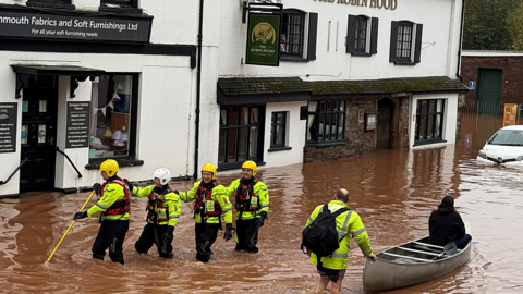 Four rescuers in hi-vis wade through flooded street as a man in a hi-vis jacket pushes a person in a canoe. There is a pub on the left of the image and a shop. They are both white buildings.
