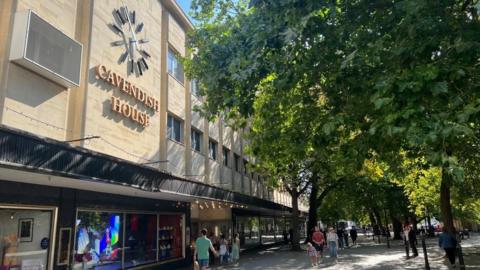 The front of Cavendish House with its name on the front of the building, a large clock, as pedestrians walk past among green trees. 