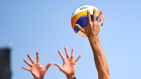 A close-up of a person hitting a volleyball and two hands trying to defend it. The sky is blue.