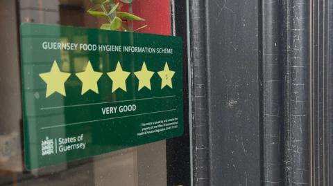 A photo of a green sticker on a window. The sticker has five stars on it and is titled 'Guernsey Food Hygiene Information Scheme'