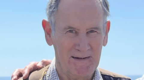 Head and shoulders photo of Richard Parker. He has grey hair and is wearing a check shirt and brown jacket. He is looking into the camera with a slight smile. Someone is standing next to him out of shot but they have their arm around him and their hand is on his shoulder.