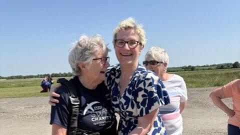 Avril Abraham is wearing a parachute harness stands on an airfield, being hugged by Sarah Allen in a patterned dress. Avril is wearing a dark T-shirt with a charity logo. In the background, a few other people stand on the gravel area, with open fields and a clear blue sky behind them.