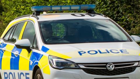 A British police car is driving along a country lane. You cannot see who is driving as the car is dark inside.