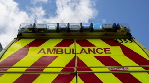 A close-up of the back of an ambulance, with diagonal red and yellow stripes and the word 'Ambulance' on the back in red. Blue lights are on the top of the vehicle and the sky is visible above the ambulance.
