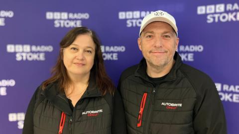 Jill and Mark Moss, wearing black Autobrite Direct branded gilets, are stood against a purple BBC Radio Stoke branded backdrop.