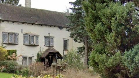 A house in the background of the photo, including three sets of windows on the first floor on the left hand side of the photo and a single window on the same floor. Trees taller than the house are also in the image.