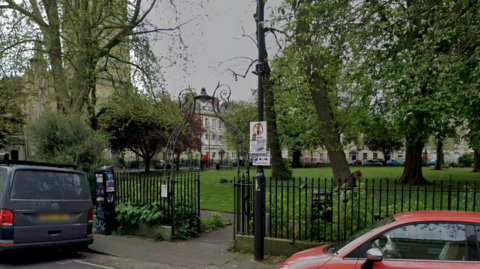 Portland Square, a small grassy park surrounded by black mteal fencing. There are several trees in the park, and a red car and grey van are parked on the road outside it.