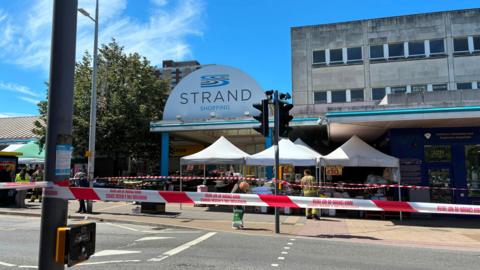 A picture of the shopping centre with a blue sky as the backdrop. Red tape is at the front of the image and cordons off the centre