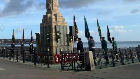 A tall war memorial on a seaside promenade, surrounded with standard bearers carrying flags for the ceremony. 