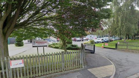 The outside of a school building, which has wooden fences and a black metal gate. There is a white sign that says Whitestone infant school in red lettering. Trees obscure the view of the school, but cars are visible in a car park behind the gates