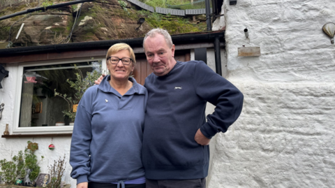 A man and woman stand side by side. The man's arm is around the woman's shoulder. She is wearing a light blue fleece and has short blonde hair and black square glasses. He is swearing a navy blue sweatshirt and has short grey hair.