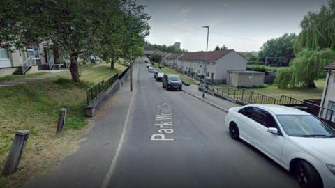 Parkwood Crescent in Leeds. A narrow road with cars parked on one side and a grass verge on the left with trees. A row of terraced houses are on the right.