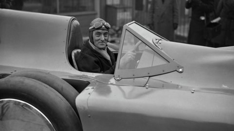 A black‑and‑white photo of a person in a vintage open race car, wearing a cap and goggles, with a few onlookers in the background.