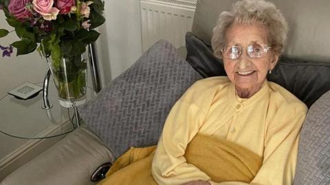 Lilian Cox is smiling and wears glasses and has a yellow cardigan. She is in a chair with a large bunch of roses on a table next to her