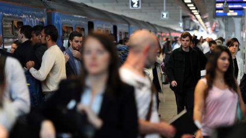 Men and women on a train platform during a busy period where commuters are making their way to work. A dark blue train is parked next to the platform.