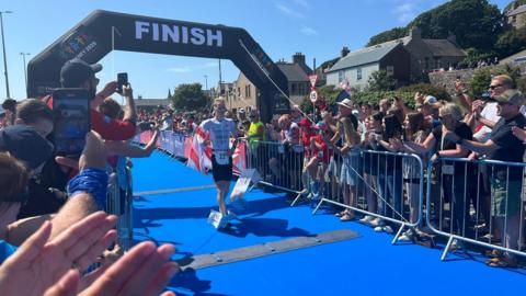 A runner crosses a blue-floored finish line beneath a large “FINISH” arch while spectators line both sides of the course, cheering and taking photos on a sunny day.