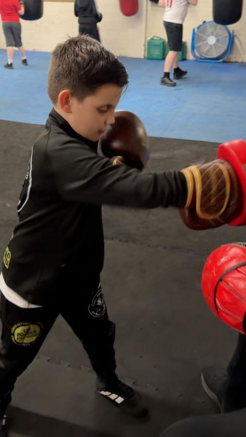 A young boy wearing black sports clothing punches punching pads as he wears black and brown boxing gloves in a gym setting