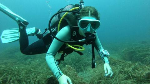 Mia is seen scuba diving near the seafloor, wearing a mask, fins, and full diving gear. Bubbles rise from her regulator as she glides over rocks and sea grass in clear blue water.