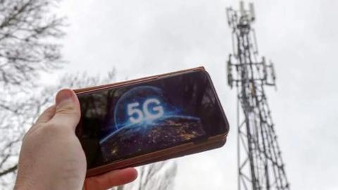 A hand holding a mobile phone with the letters 5G on it, with a mobile phone mast in the background