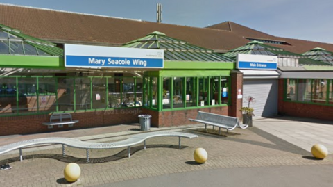 The entrance to Royal South Hants Hospital consists of a main brown tiled roof, with a green conservatory-style glass structure at the front with four separate triangular roofs. One large winding metal bench and one back-to-back bench sit  on the pavement in front.