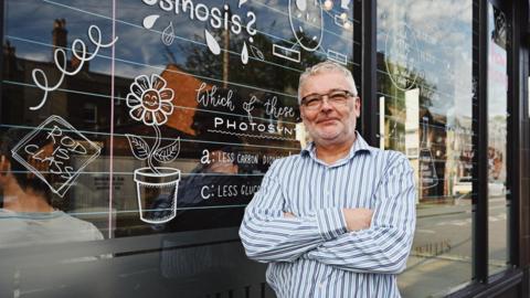 Willi's Cafe owner Rod Davis stands in front of a window covered in writing and doodles, including a drawing of a plant pot. He is wearing glasses and a blue/grey striped shirt. There are customers inside the cafe and street reflections in the glass.