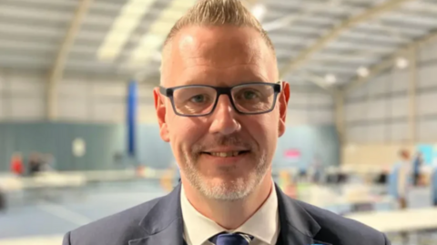 A close-up image of a man with short blonde hair who is wearing dark-framed glasses, a cream-coloured shirt and blue tie and a grey suit. The room behind him is blurred.