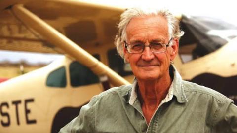 Iain Douglas-Hamilton stands in front of a plane wearing glasses and a grey collared shirt