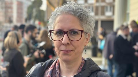A head-and-shoulders portrait of Rivka Gottlieb. She has grey hair, large black glasses and a red-and-pink patterned scarf. She is pictured with a crowd of people behind her outside the inquiry room in West London.