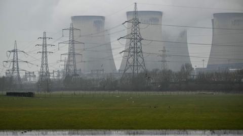 A power station pictured from across a field and behind some electric pylons.