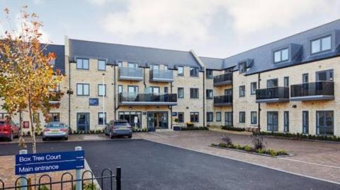 An exterior view of Box Tree Court in Boston Spa. It is a three-storey building built in light brick, with some balconies and a grey roof. Cars are parked outside the building, and there is a sign saying, "Box Tree Court, Main entrance" in the foreground.