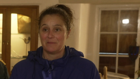 The image shows a person standing indoors wearing a dark blue hooded jacket. Behind them is a wooden display cabinet with glass doors containing various household items. To the right is a window with multiple panes.
