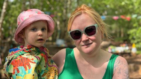 A woman in her early thirties with red hair wearing green sunglasses holding her infant daughter. They are in a woodland area with green leaves on trees visible behind them.