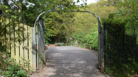 Metal gates at one of the entrances to Saltburn's Valley Gardens. They are open, leading on to a path with trees in the background. Wooden fences stand either side of the gates.