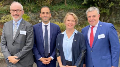 Four people who are officials from France and Guernsey are pictured standing shoulder to shoulder and they are all smiling at the camera. There are three men and one woman. The men are all wearing suits. The woman is wearing a blue shirt with a dark blue cardigan.
