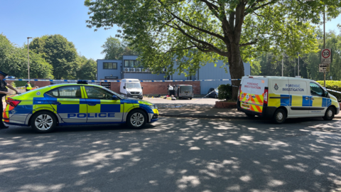 Police tape with parked vans being inspected by officers behind the cordon