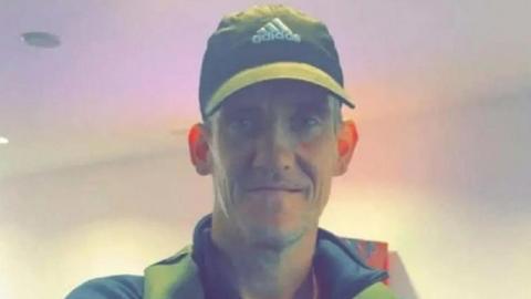 Dale Stogden, wearing an Adidas cap and a dark shirt with a large collar. He is stood in front of a pale background and is smiling at the camera.